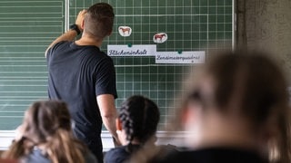 Ein Lehrer macht im Unterricht einer vierten Klasse einer Grundschule einen Tafelanschrieb, während an der Tafel Zettel mit der Aufschrift "Flächeninhalt" und "Zentimeterquadrat" hängen. 