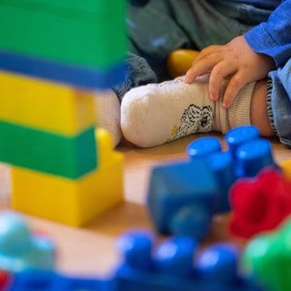 Ein Kleinkind sitzt vor einem Turm aus bunten Bauklötzen. Zu sehen ist sein Fuß und seine Hand. 