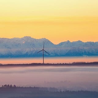 Drei Windräder ragen am Morgen kurz nach Sonnenaufgang aus der Nebeldecke, während im Hintergrund durch den Föhn die über einhundert Kilometer entfernten Alpen zu sehen sind.