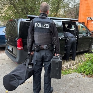 Ein Polizist steht bei einer Durchsuchung mit einer Tasche und einem Koffer neben einem Wagen.