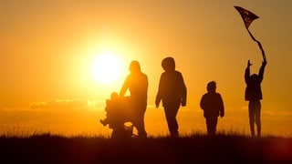 Eine Familie mit Kinderwagen und einem Drachen an der Schnur geht bei Sonnenuntergang über einen Weg durch die Salzwiesen im Watt bei Westerhever (Symbolbild).