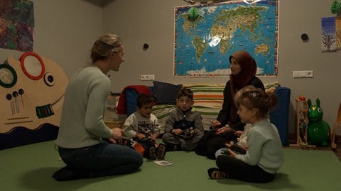 Mehrere Kinder sitzen mit zwei Betreuerinnen auf dem Boden und singen gemeinsam.