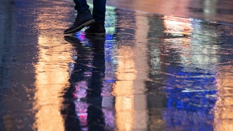 Ein Mann geht im Regen über eine gepflasterte Fläche, in der sich Lichter der umliegenden Geschäfte spiegeln.