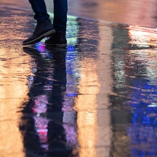 Ein Mann geht im Regen über eine gepflasterte Fläche, in der sich Lichter der umliegenden Geschäfte spiegeln.