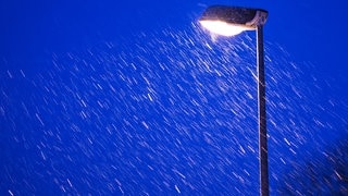 Leichter Schneefall am Abend. Eine Straßenlaterne leuchtet.