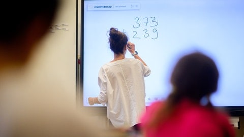 Eine Lehrerin schreibt eine Mathematikaufgabe auf eine digitale Schultafel im Klassenraum einer 4. Klasse einer Grundschule.