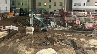 Ein Bagger steht auf einer Baustelle hinter einer Bombe, die mit einer schwarzen Plastikplane abgedeckt ist. Wegen eines Bombenfunds müssen einige Menschen in Teilen der Ulmer Innenstadt ihre Wohnungen verlassen. Bauarbeiter hätten am Nachmittag eine rund 250 Kilogramm schwere Fliegerbombe auf einer Baustelle gefunden, teilte die Polizei mit. 