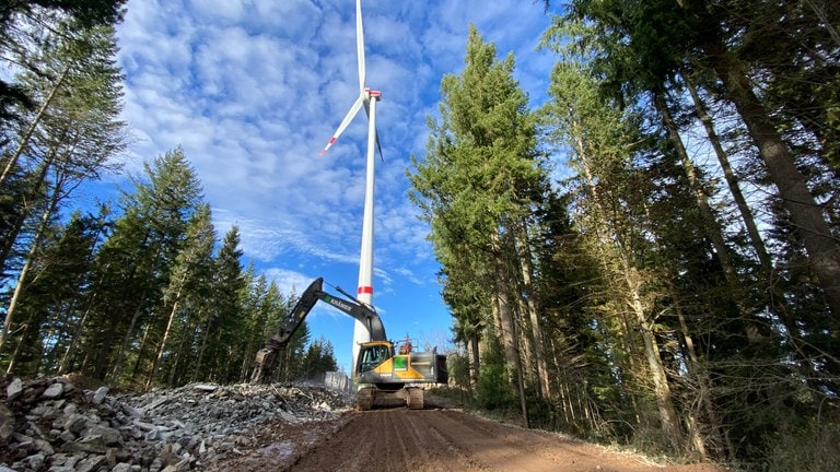 Ein Bagger steht vor einem Windrad im Wald. Neben dem Windrad liegen Trümmerteile eines Windrads.