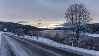 Die aufgehende Sonne beleuchtet den Himmel über dem Schluchsee während im Vordergrund eine schneebedeckte Straße zu sehen ist. Die Schneefallgrenze liegt derzeit bei 800 Metern.