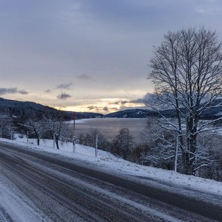 Die aufgehende Sonne beleuchtet den Himmel über dem Schluchsee während im Vordergrund eine schneebedeckte Straße zu sehen ist. Die Schneefallgrenze liegt derzeit bei 800 Metern.