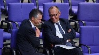 Thorsten Frei (links) und Friedrich Merz sitzen im Bundestag und unerhalten sich. Beide lächeln.