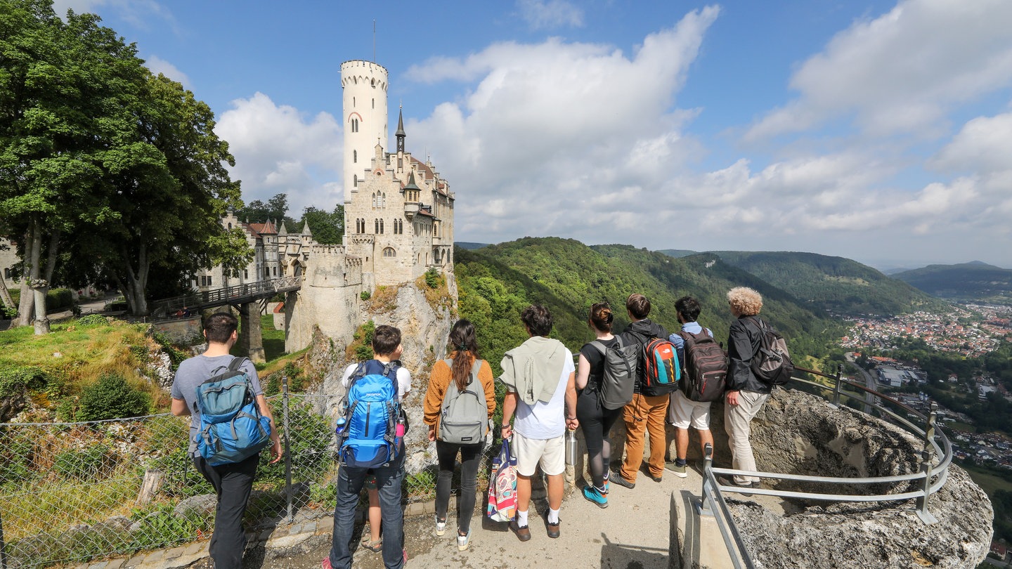 Touristen stehen auf einer Aussichtsplattform und blicken auf das Schloss Lichtenstein auf der Schwäbischen Alb. Im Sommer 2024 waren die Übernachungszahlen in BW auf einem neuen Höchststand.