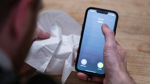 Ein Mann hält ein Taschentuch in einer Hand und ein Telefon mit dem Schriftzug "Arzt" in der Anderen. 