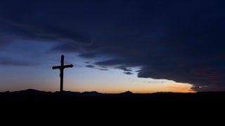 (Symbolbild) Eine Wolkenfront zieht hinter einem Wegkreuz über die im Sonnenuntergang liegende Berglandschaft.