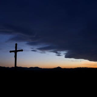 (Symbolbild) Eine Wolkenfront zieht hinter einem Wegkreuz über die im Sonnenuntergang liegende Berglandschaft.