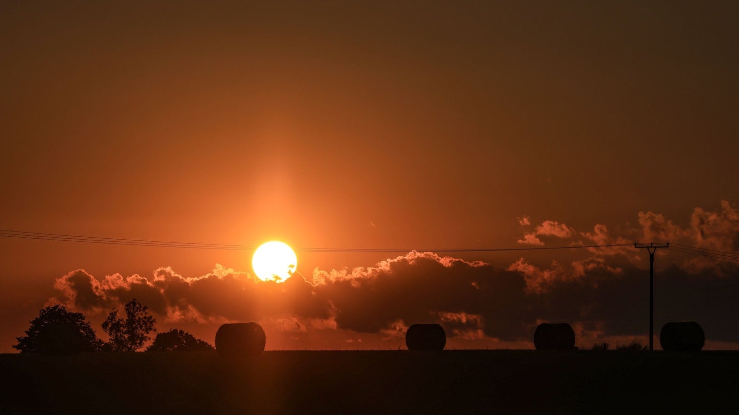 Strohballen liegen auf einem Feld, während die Sonne am Horizont untergeht.