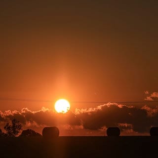 Strohballen liegen auf einem Feld, während die Sonne am Horizont untergeht.