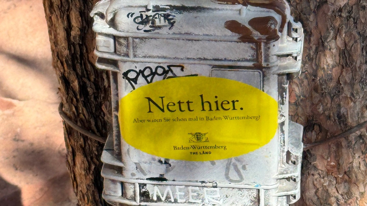 Auf einem Kasten an einem Baum klebt ein Aufkleber unter anderem mit der Aufschrift 