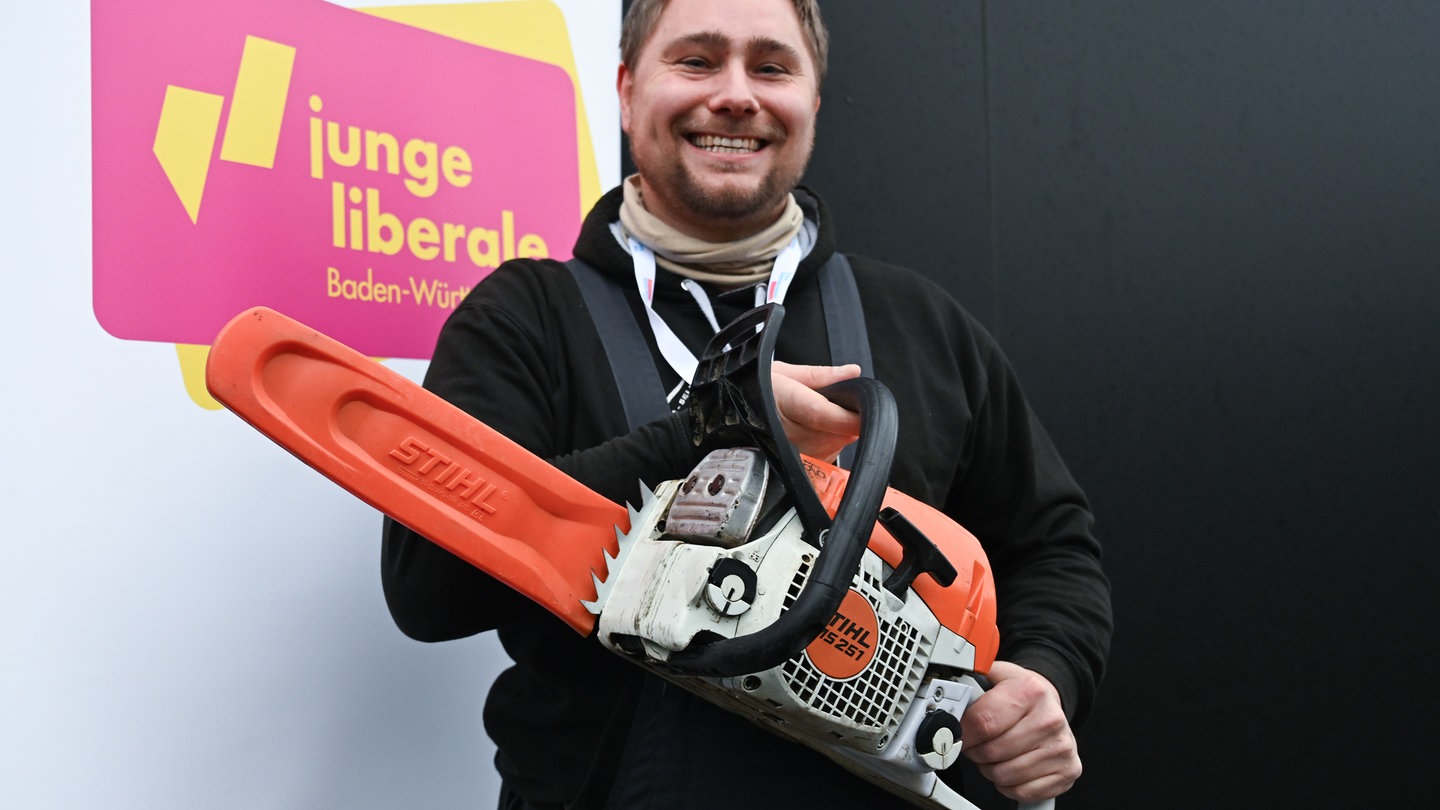 Mark Hohensee, Landesvorsitzender der Jungen Liberalen, der Jugendorganisation der FDP, hält vor dem Eingang der Schwabenlandhalle vor dem Beginn des Landesparteitags der Südwest-FDP eine Motorsäge in der Hand. Er will damit für einen Bürokratieabbau mit der Kettensäge demonstrieren.