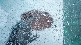 Eine Passantin geht mit ihrem Regenschirm an einer Scheibe mit Regentropfen in der Innenstadt vorbei. Viele Menschen geben an, dass sie Wetterveränderungen spüren können. Sie leiden beispielsweise an Kopfschmerzen oder Schwindel. 