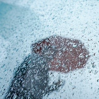 Eine Passantin geht mit ihrem Regenschirm an einer Scheibe mit Regentropfen in der Innenstadt vorbei. Viele Menschen geben an, dass sie Wetterveränderungen spüren können. Sie leiden beispielsweise an Kopfschmerzen oder Schwindel. 