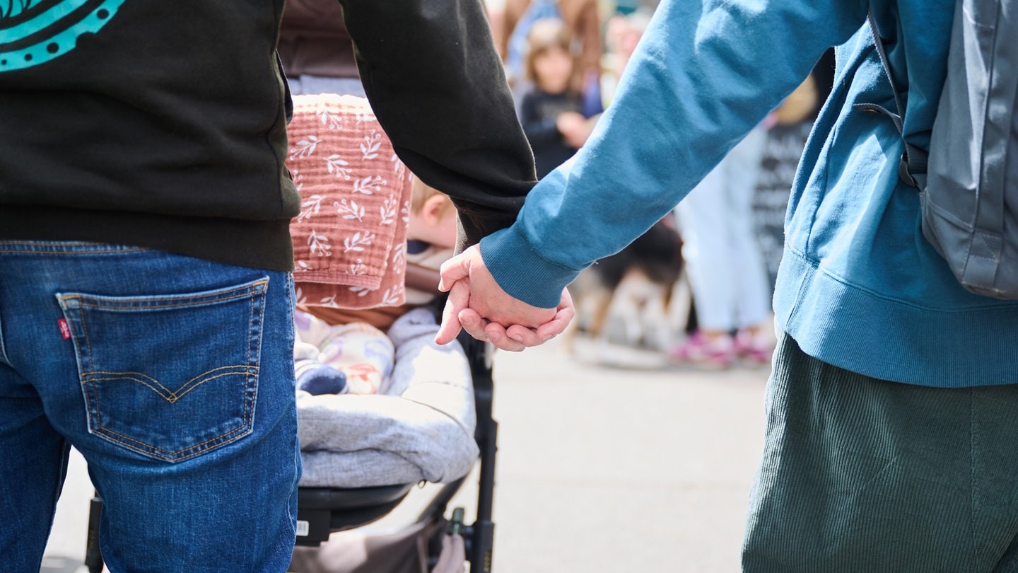 Eltern halten Händchen, während sie mit ihrem Baby im Kinderwagen über einen Markt gehen.