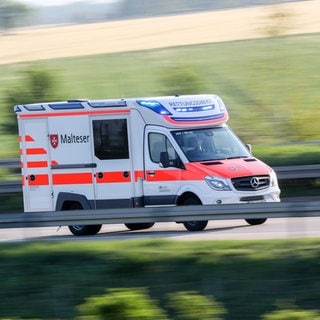 Symbolbild | Ein Rettungswagen der Malteser fährt mit Blaulicht über eine Straße, im Hintergrund sind verschommen Felder zu sehen. 