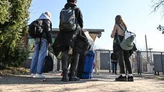 Drei Personen mit Rucksäcken, Koffern und Jacken stehen vor einem Gitterzaun und einem Häuschen, im Hintergrund eine Hecke und blauer Himmel. Junge Geflüchtete stehen 2022 vor der Landeserstaufnahmeeinrichtung in Sigmaringen in Baden-Württemberg.