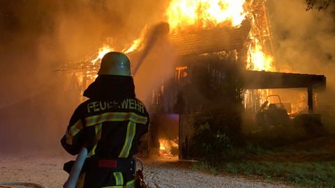Ein Feuerwehrmann steht mit einem Schlauch vor einer brennenden Scheune und löscht.