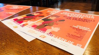 Auf einem Tisch liegen meherere orangefarbene Flyer aufeinander für die Aktionstage Gewalt gegen Frauen.