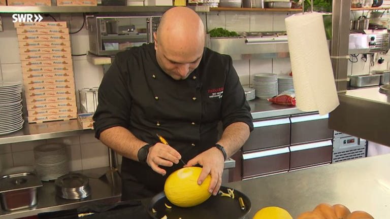 Guiseppe Pasarelli steht an einem Küchentisch und schnitzt mit einem Messer ein Muster in eine Melone.