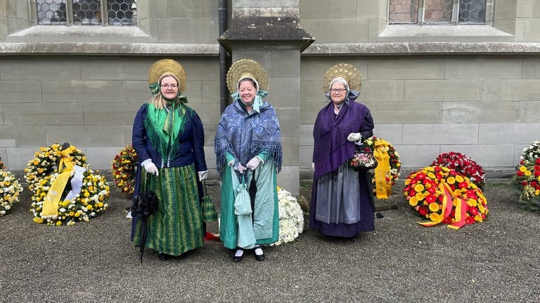 Drei Frauen einer Trachtengruppe, die zur Trauerfeier nach Salem gekommen sind, im Hintergrund zahlreiche Trauerkränze.