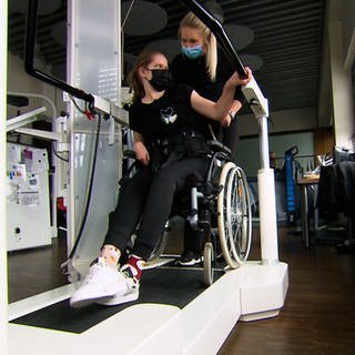 Frau im Rollstuhl wird von Therapeutin auf ein Therapiegerät geschoben.
