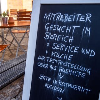 Ein Schild steht vor einer Gaststätte. Darauf steht, dass Mitarbeiter gesucht werden für Service und Küche.