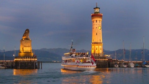 Ein Schiff fährt an den Wahrzeichen der Stadt Lindau am Bodensee, dem Bayerischen Löwen und dem Leuchtturm, aus dem Hafen.