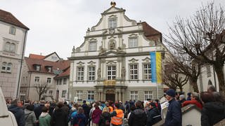 Viele Menschen stehen vor einem Rathaus, vor dem Gebäude ist die urkainische Nationalflagge gehisst.