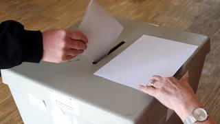 Eine Hand wirft einen Stimmzettel in eine Wahlurne.