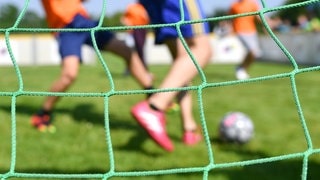 Jugendliche spielen Fußball auf einem Fußballplatz. Im Vordergrund ist das Netz des Fußballtors zu sehen.