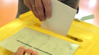 Eine Hand führt einen Stimmzettel in eine Wahlurne.