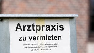 Ein Schild mit der Aufschrift "Arztpraxis zu vermieten" steht vor einem Gebäude.