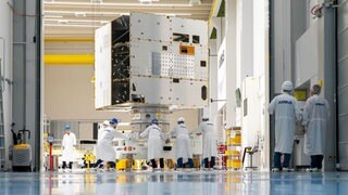 Ein Satellit steht in einer Halle, mehrere Menschen in weißer Arbeitskleidung stehen ebenfalls in der Halle.