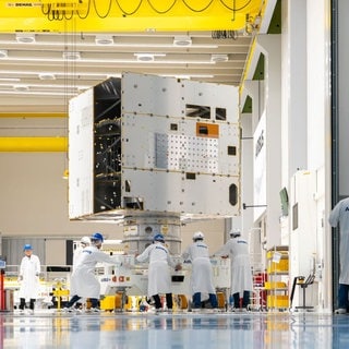 Ein Satellit steht in einer Halle, mehrere Menschen in weißer Arbeitskleidung stehen ebenfalls in der Halle.