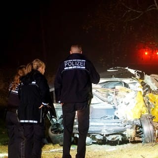 Einsatzkräfte der Polizei stehen vor dem Wrack eines verunfallten Autos. 