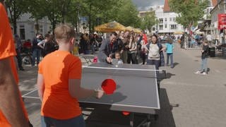 An einer Tischtennisplatte spielen zwei Spieler, während hunderte in der Schlange stehen und warten bis sie drankommen. 