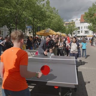 An einer Tischtennisplatte spielen zwei Spieler, während hunderte in der Schlange stehen und warten bis sie drankommen. 