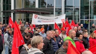 Menschen stehen in Friedrichshafen und streiken. Einige haben Fahnen der IG Metall dabei.