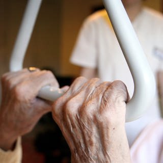 Eine Pflegehausbewohnerin in einem Seniorenzentrum hält sich in ihrem Bett an einem Haltegriff fest, im Hintergrund steht eine Pflegekraft.