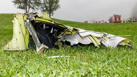 Flugzeugwrackteil auf einer Wiese. Rettungsfahrzeuge stehen im Hintergrund.