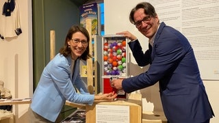 Bettina Gräfin Bernadotte und ihr Bruder Björn Graf Bernadotte stehen an einer Art Kaugummi-Automaten, in dem sich Kugeln mit Zetteln befinden. Auf den Zetteln stehen Witze. Eine Kindheitserinnerung der beiden.