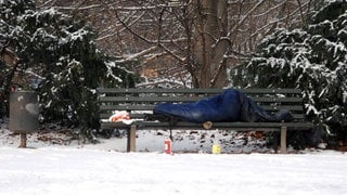 Ein Obdachloser liegt im Schlafsack auf einer Parkbank im Schnee.